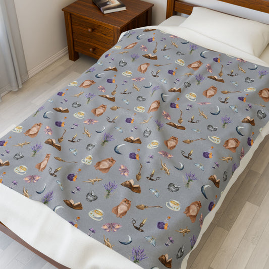cozy bed decorated with printed throw featuring witchy aesthetic cats, brooms, cauldrons, herbs, potions, moths, and mushrooms